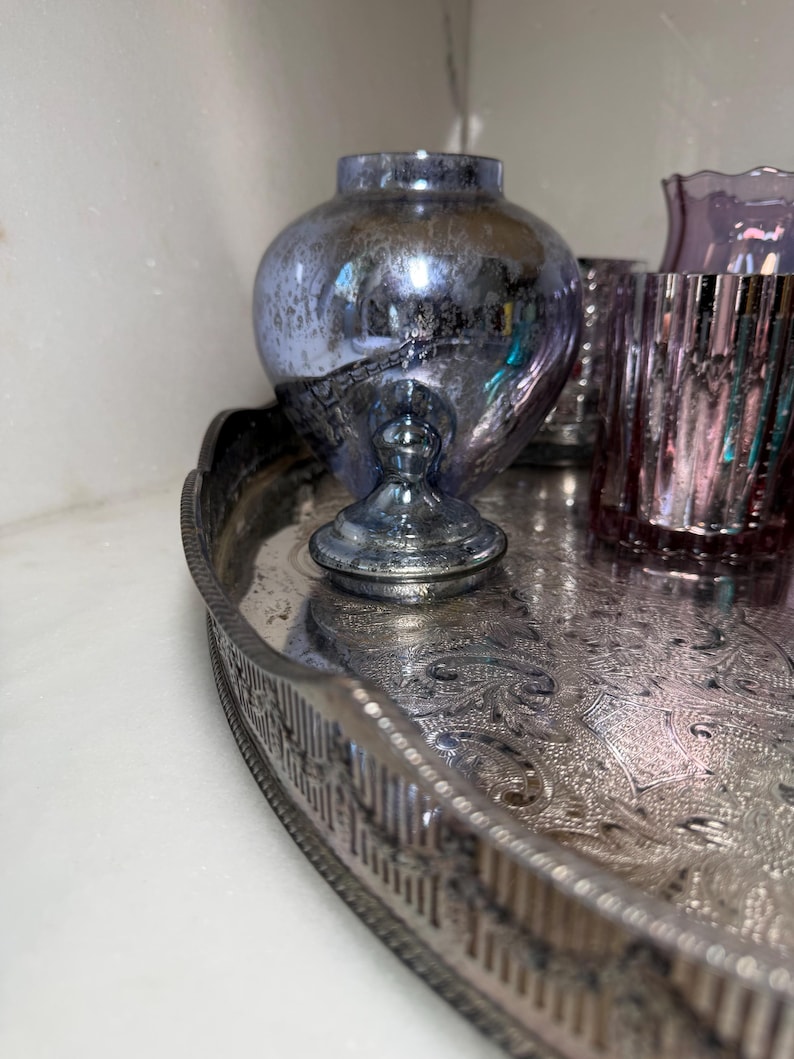 Peut inclure: Un plateau argent&eacute; orn&eacute; contient une collection d'objets en verre d&eacute;coratifs. Un grand vase bleu&acirc;tre de forme bulbeuse repose sur un pi&eacute;destal. Plusieurs verres roses et violets sont &eacute;galement sur le plateau.