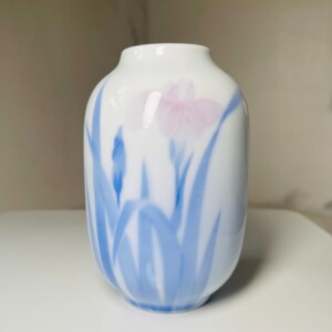 May include: White ceramic vase with a painted design of a pink iris flower and blue leaves. The vase has a rounded body and a narrow neck. The floral design is delicate and elegant.