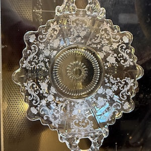 May include: Clear glass serving dish with a floral and scroll design. The dish has a scalloped edge and two handles. The center of the dish has a circular pattern. The floral design is etched into the glass.