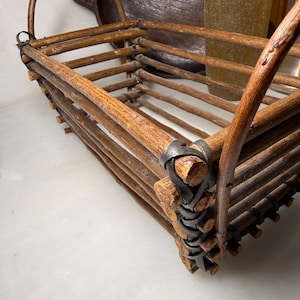 May include: A rectangular basket constructed from brown wooden twigs and branches, featuring two arched handles on the sides. The structure is held together by dark, flexible binding strips wrapped around the corners and along the base frame.