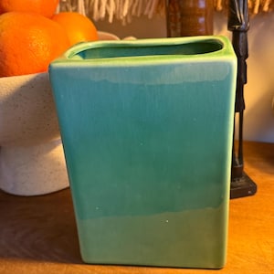 May include: A square, turquoise-green ceramic vase with a slightly tapered shape. The vase has a glossy finish and a light green rim. The background includes oranges, a ceramic bowl, and decorative items.