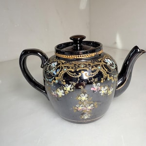 May include: Black ceramic teapot with lid, featuring floral and gold accents. The teapot has a curved handle and spout. Delicate floral patterns in pink, yellow, and blue adorn the body, with gold trim around the lid.