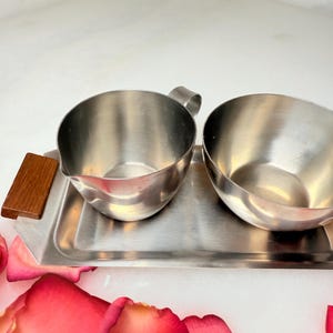 May include: A stainless steel creamer and sugar bowl set on a rectangular tray with a wooden handle. The creamer has a handle and a spout. The sugar bowl is round. The set is on a white surface with pink rose petals.