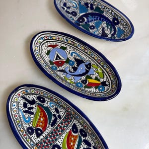 Könnte beinhalten: Drei ovale Keramikschalen mit handbemalten Fischmotiven in leuchtenden Blau-, Rot-, Gelb- und Grüntönen. Jede Schale ist mit komplexen schwarzen Punktmustern und einem kräftigen blauen Rand verziert, was ein markantes aquatisches Design ergibt.
