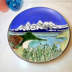May include: A round decorative plate featuring a colorful enamel landscape scene. The design depicts a blue sky, white snow-capped mountains, a green hill, a light blue body of water, a sandy shore, and green cacti in the foreground.