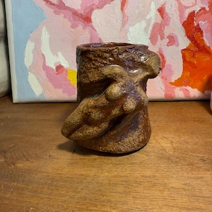 May include: A brown ceramic vase with a textured exterior and a sculpted figure of a person on the side. The vase has a cylindrical shape and is sitting on a wooden surface.
