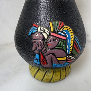 May include: A black ceramic vase with a textured surface and a yellow base and rim. The vase features a colorful relief of two figures with elaborate headdresses. The figures are in shades of blue, red, yellow, and purple.