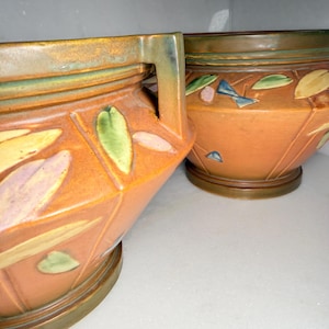 May include: Two terracotta-colored ceramic planters featuring a geometric faceted design. Each planter is decorated with raised, multi-colored leaf motifs in shades of green, yellow, and purple, and includes a dark green rim and base.