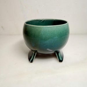 May include: A round, deep green ceramic bowl with a smooth, glossy finish. The bowl features a wide, open top and is supported by three short, sturdy, integrated feet that elevate the base slightly above the surface it rests upon.