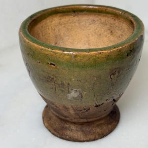 May include: A small, antique-style ceramic bowl with a rounded shape and a pedestal base. The bowl features a distressed, textured finish with a green rim and a brown body. The interior is a lighter brown color.