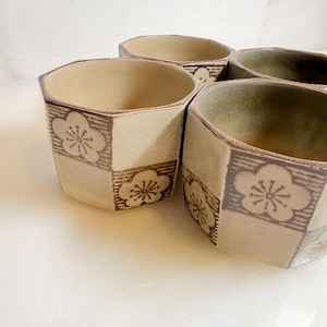 May include: A set of four ceramic cups with a hexagonal shape and a cream-coloured glaze. Each cup features a repeating pattern of brown square blocks containing a stylised five-petalled flower design. The interior of the cups has a smooth finish.