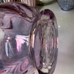 Peut inclure: Gros plan sur un vase en verre violet de forme arrondie et &agrave; bord &eacute;vas&eacute;. Le verre pr&eacute;sente un effet tourbillonnant et iris&eacute;, avec des nuances de rose et de lavande. D'autres objets en verre sont visibles en arri&egrave;re-plan.