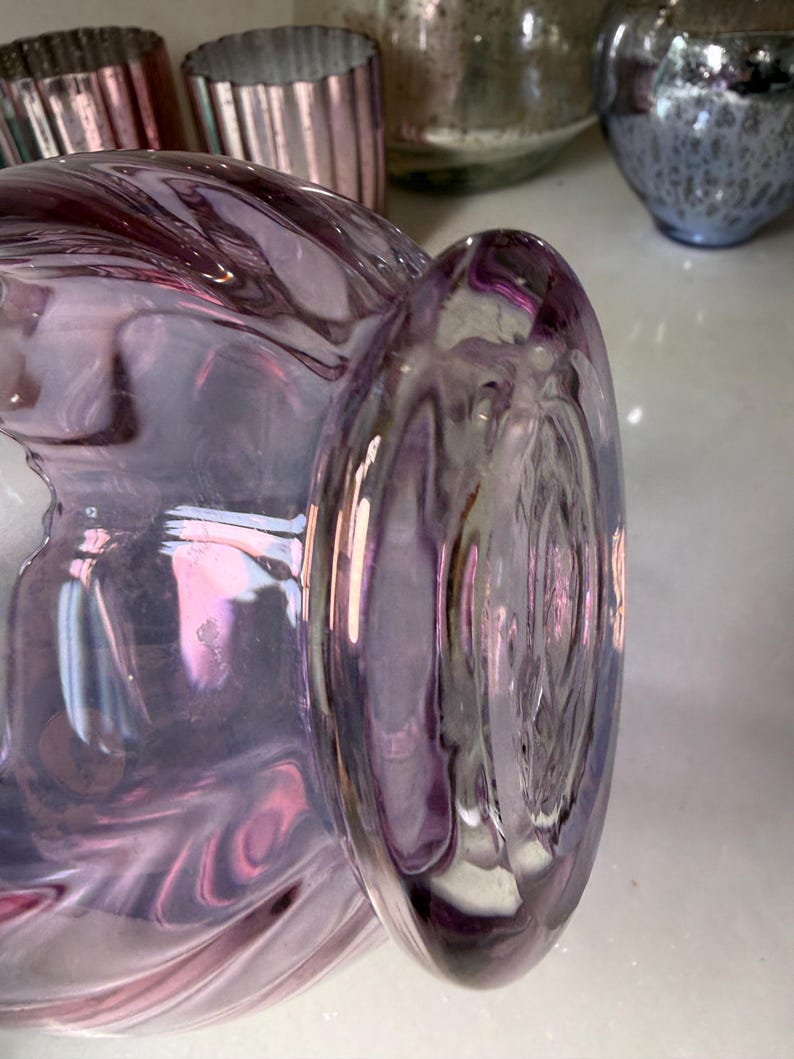 Peut inclure: Gros plan sur un vase en verre violet de forme arrondie et &agrave; bord &eacute;vas&eacute;. Le verre pr&eacute;sente un effet tourbillonnant et iris&eacute;, avec des nuances de rose et de lavande. D'autres objets en verre sont visibles en arri&egrave;re-plan.