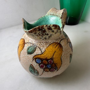 May include: A round ceramic pitcher with a textured beige surface featuring hand-painted yellow flowers with multi-colored centers and green leaves. The interior of the pitcher is glazed in a bright turquoise color, and the rim has a brown speckled finish.