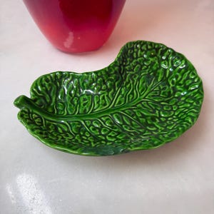 Puede incluir: Un plato de cerámica de color verde brillante con forma de hoja de col texturizada. El plato presenta intrincados detalles de nervaduras en relieve y una superficie arrugada que imita la apariencia de una hoja de verdura real con un tallo.