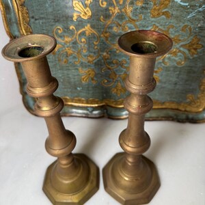 May include: Two antique brass candlesticks with a hexagonal base and a slender, curved body. The candlesticks have a tarnished, aged appearance, suggesting they are vintage. The background features a teal tray with gold floral detailing.