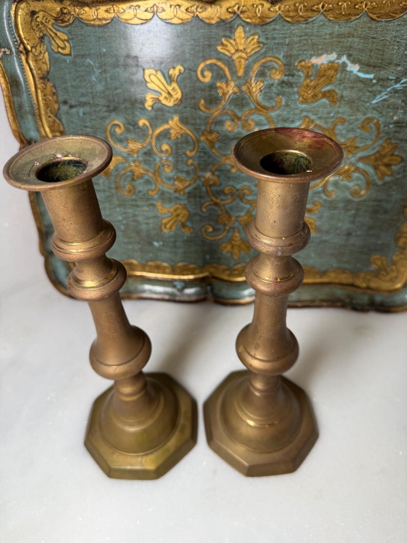 May include: Two antique brass candlesticks with a hexagonal base and a slender, curved body. The candlesticks have a tarnished, aged appearance, suggesting they are vintage. The background features a teal tray with gold floral detailing.