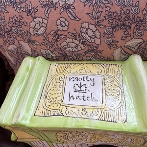 May include: A lime green ceramic birdhouse with the words "molly hatch" in a white rectangle. The birdhouse has a decorative design with yellow and black accents. The background is a pink floral pattern.