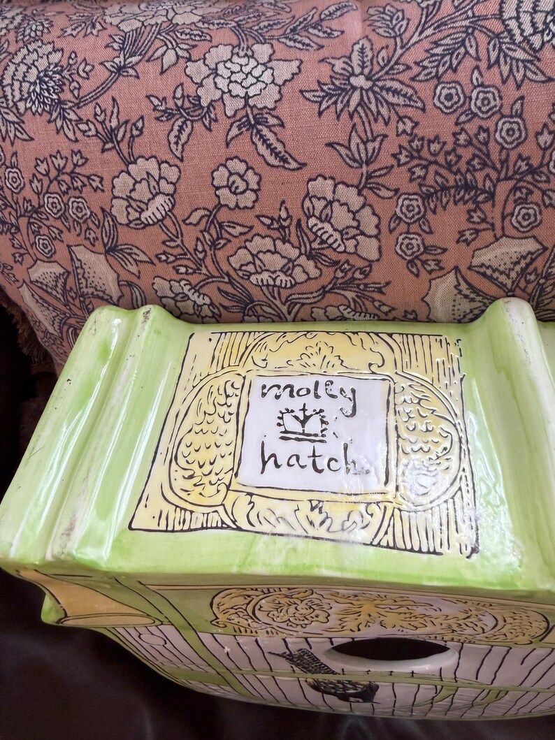 May include: A lime green ceramic birdhouse with the words "molly hatch" in a white rectangle. The birdhouse has a decorative design with yellow and black accents. The background is a pink floral pattern.