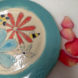 May include: A handmade ceramic plate with a turquoise rim and a cream-colored center. The plate features hand-painted flowers in red, blue, and yellow, along with a small bee. Pink rose petals are scattered nearby.