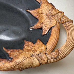 May include: A black ceramic serving tray with a brown, leaf-themed handle. The handle features detailed leaf and vine designs. The tray's rim and handle are a warm brown, contrasting with the black interior.