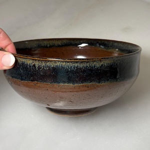 May include: A handmade ceramic bowl with a dark brown interior and a black and brown exterior. The bowl has a glossy finish and a slightly flared rim. The bowl is approximately 6 inches in diameter and 3 inches tall.
