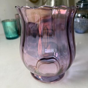 Peut inclure: Un photophore d&eacute;coratif en verre violet avec un bord festonn&eacute;. Le verre est translucide, avec des reflets roses et iris&eacute;s. Le support a un corps arrondi et une petite base circulaire. D'autres photophores sont visibles en arri&egrave;re-plan.