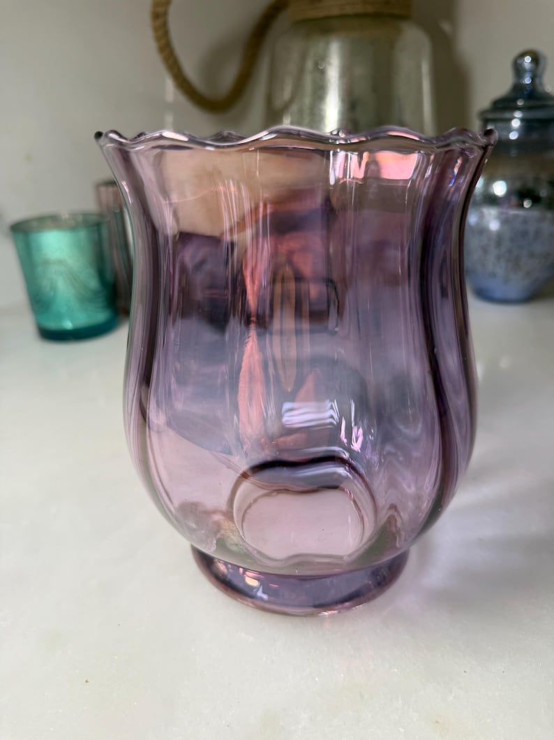 Peut inclure: Un photophore d&eacute;coratif en verre violet avec un bord festonn&eacute;. Le verre est translucide, avec des reflets roses et iris&eacute;s. Le support a un corps arrondi et une petite base circulaire. D'autres photophores sont visibles en arri&egrave;re-plan.
