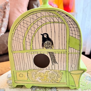 May include: A ceramic birdcage-shaped decorative piece in a vibrant lime green. The cage features a black and white bird illustration inside, with black bars and a circular opening. The base has a yellow floral design.