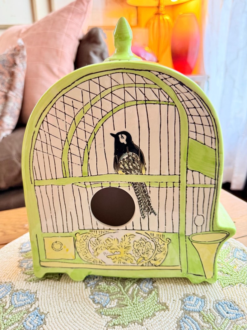May include: A ceramic birdcage-shaped decorative piece in a vibrant lime green. The cage features a black and white bird illustration inside, with black bars and a circular opening. The base has a yellow floral design.