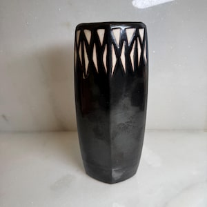 May include: A cylindrical ceramic vase with a glossy black finish. The upper portion features a repeating geometric pattern of white diamond-shaped cutouts or inlays that contrast against the dark surface, creating a sharp visual border.