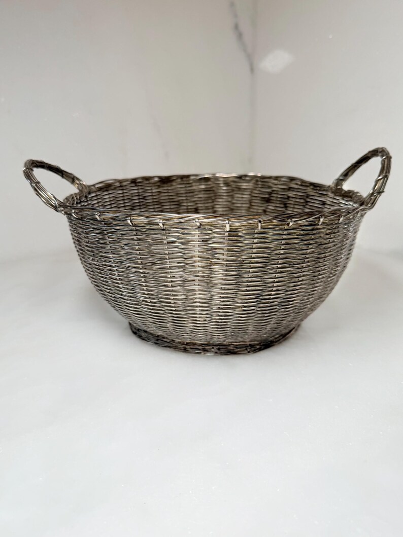 Puede incluir: Una cesta redonda de alambre plateado con dos asas arqueadas. La cesta tiene un dise&ntilde;o tejido y una forma ligeramente acampanada. La cesta est&aacute; vac&iacute;a y se encuentra sobre una superficie blanca.