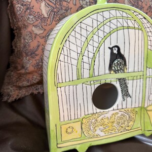 May include: A ceramic birdhouse shaped like a cage, painted in white, black, and lime green. The birdhouse features a black and white bird perched inside. The base has a yellow and gold decorative design.