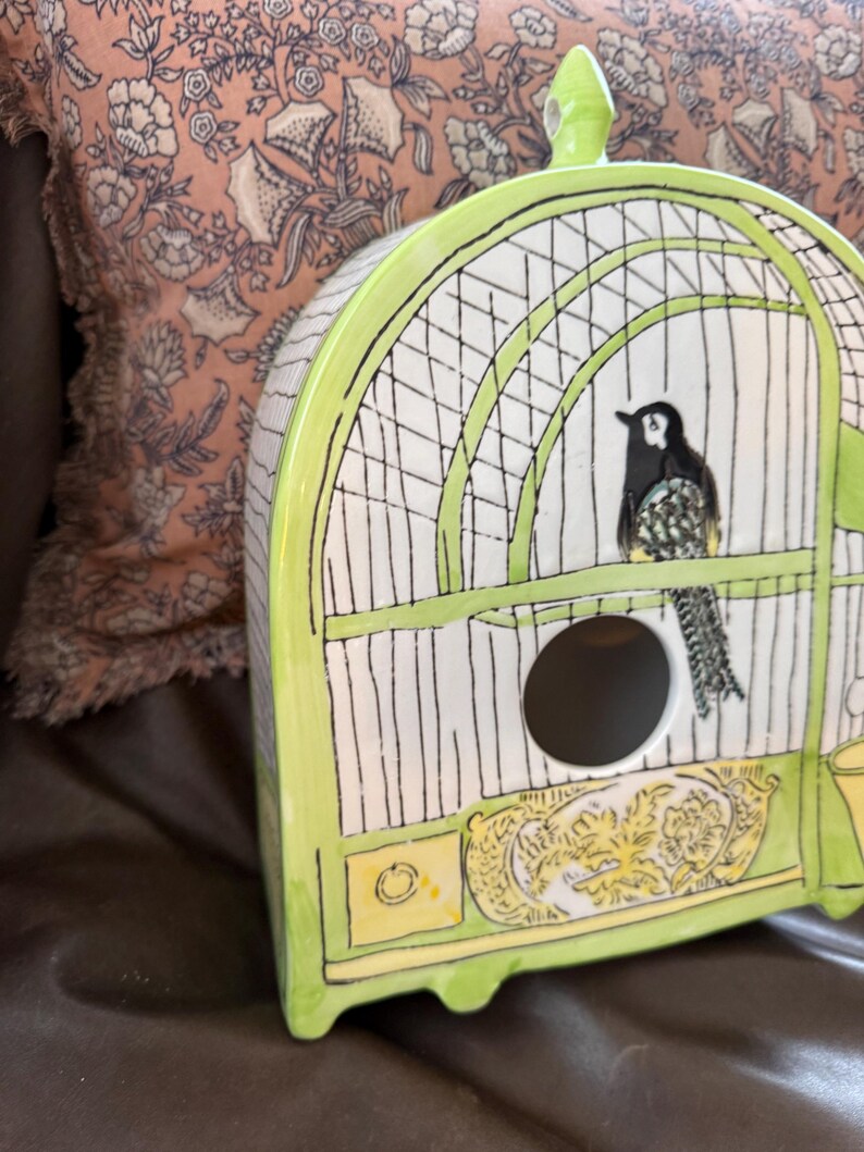 May include: A ceramic birdhouse shaped like a cage, painted in white, black, and lime green. The birdhouse features a black and white bird perched inside. The base has a yellow and gold decorative design.