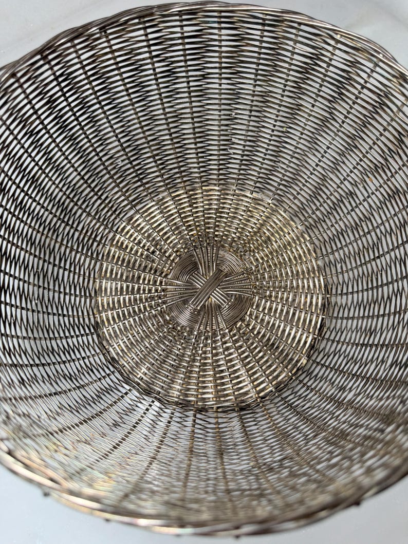 Puede incluir: Una cesta de alambre tejido de color plateado con forma redondeada. El dise&ntilde;o de la cesta presenta una serie de c&iacute;rculos conc&eacute;ntricos, creando una superficie texturizada. El interior de la cesta es visible, mostrando el intrincado patr&oacute;n de tejido.