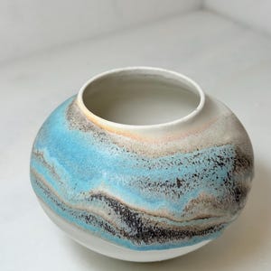 May include: A round ceramic vase with a wide opening. The vase features a blend of colors, including light blue, beige, and black, creating a layered, marbled effect. The base is white, and the interior is a light beige.