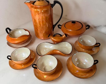 May include: A tea set featuring an iridescent orange glaze with black trim. The set includes a tall teapot, a lidded sugar bowl, a creamer, a long divided dish, and four teacups with matching saucers, all displaying a glossy, reflective finish.