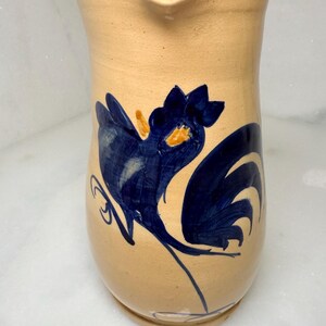 Op de afbeelding: Tan keramische kan met een blauw hanenontwerp. De haan is gestileerd geschilderd met een donkerblauwe body en staart, en oranje accenten. De kan heeft een gebogen handvat en een schenktuit.