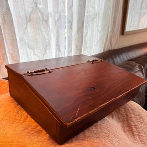 May include: A vintage wooden writing box with a hinged lid. The box is stained a deep reddish-brown color and shows signs of wear. The hinges are made of metal. The box is resting on a textured, light-colored fabric.