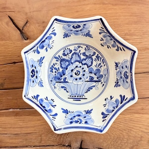May include: A white ceramic serving dish with a blue floral design. The dish has a unique octagonal shape with a scalloped edge. The center features a blue floral arrangement in a basket. Decorative and functional.