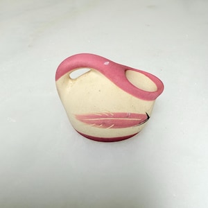 May include: A small, cream-colored ceramic pitcher with a pink handle and rim. A pink feather design is painted on the side. The pitcher has a unique, curved handle and a rounded base. The overall design is simple and elegant.