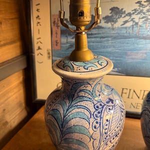 May include: A ceramic table lamp with a brass neck and a white base. The lamp features a blue and white floral pattern. The lamp is turned on and the cord is visible.