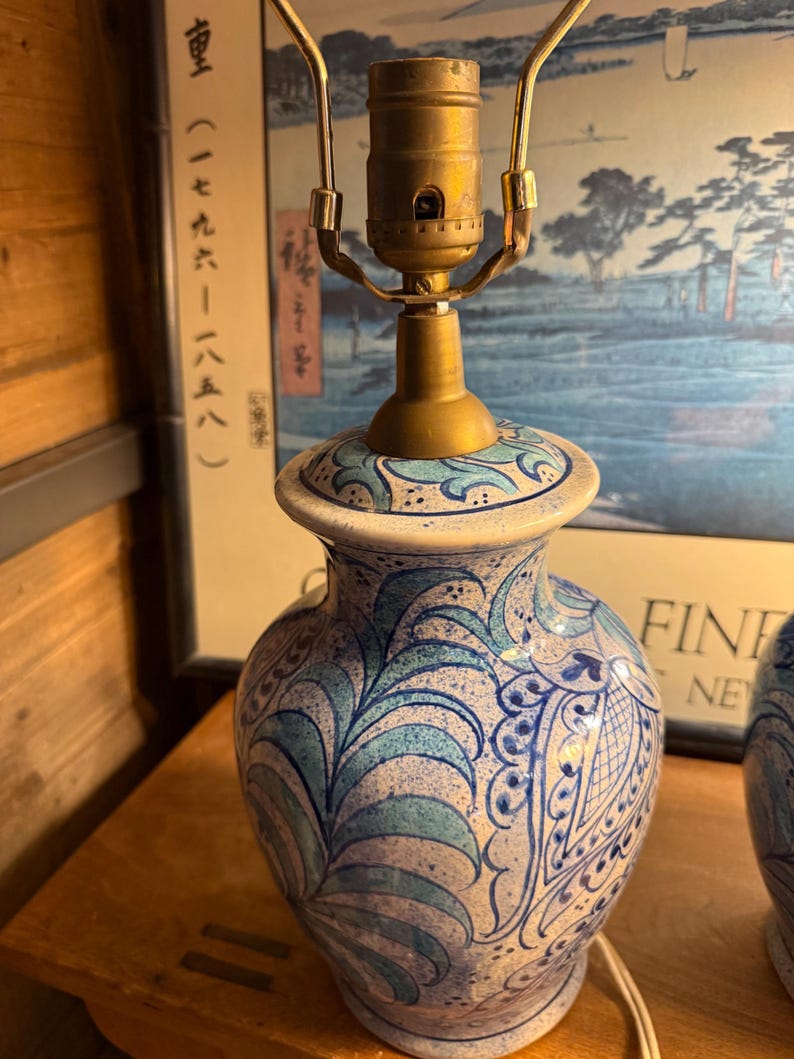 May include: A ceramic table lamp with a brass neck and a white base. The lamp features a blue and white floral pattern. The lamp is turned on and the cord is visible.
