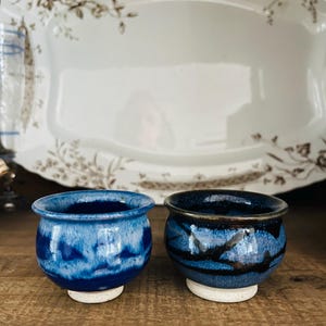 May include: Two small, handmade ceramic bowls with a blue glaze. The bowls have a rounded shape and sit on small, unglazed bases. The background features a white serving platter with a floral design.