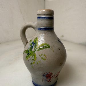 May include: A gray ceramic jug with a handle and a small spout. The jug is decorated with painted floral designs in green, blue, and red. Blue bands encircle the neck and base of the jug.