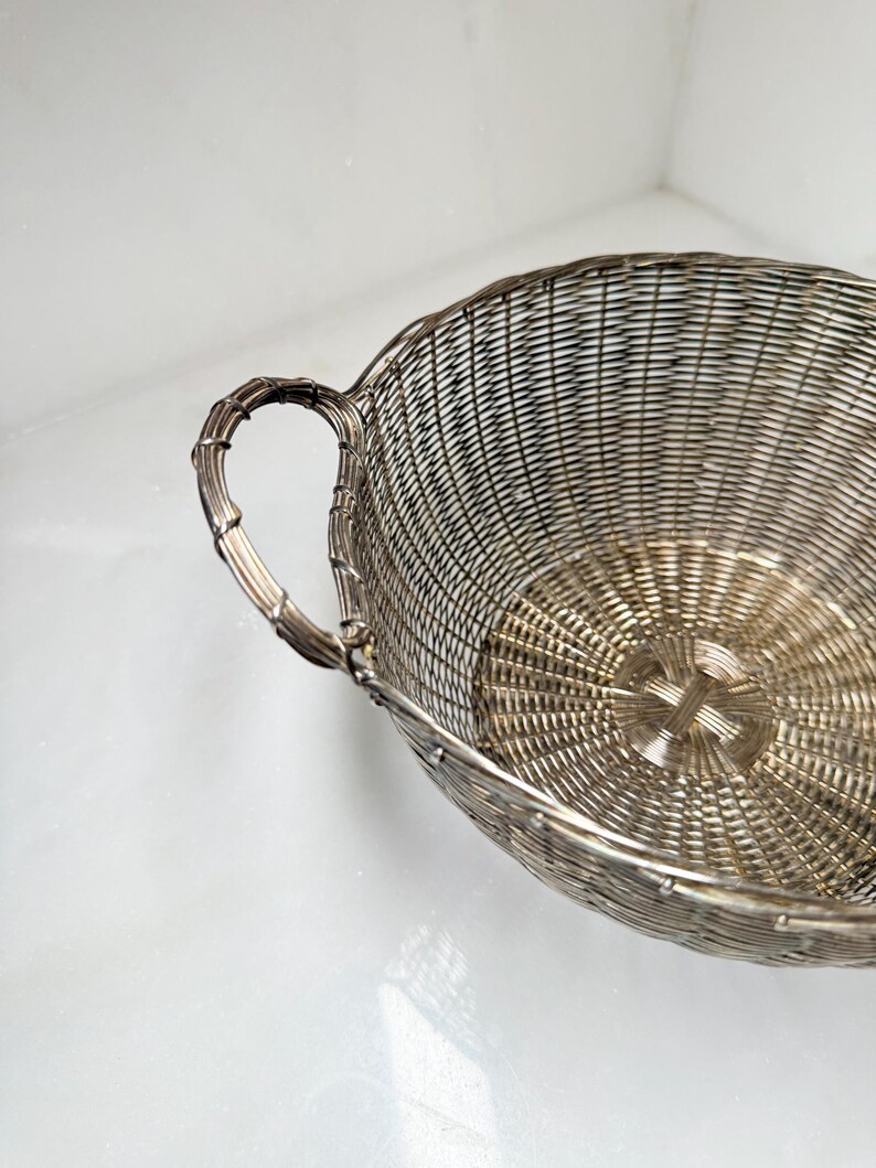 Puede incluir: Una cesta de alambre plateado con un dise&ntilde;o tejido. La cesta tiene un asa con textura de bamb&uacute;. El interior de la cesta tiene un patr&oacute;n circular que irradia desde el centro. La cesta est&aacute; sobre un fondo blanco.