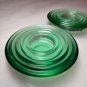 Pode incluir: Dois castiçais de vidro verde com um design de anéis concêntricos. Os castiçais têm uma forma circular com uma abertura central para uma vela. O vidro tem um acabamento brilhante e uma cor verde vibrante.