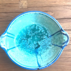 May include: A round, turquoise ceramic bowl with a textured, crystalline pattern in the center. The bowl has a blue rim and two handles. The bowl is on a wooden surface.