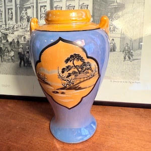 May include: A ceramic vase with a light blue body, orange rim, and handles. The vase features an orange and black landscape design. The vase is approximately 8 inches tall.