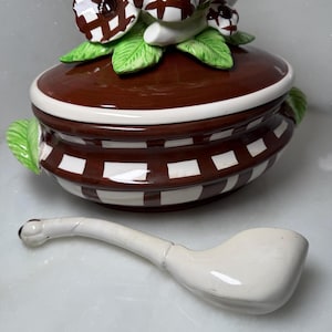 May include: A decorative ceramic serving dish with a lid, featuring a brown and white checkered pattern. The lid is adorned with floral accents in brown and white, with green leaves. A white ceramic pipe rests beside the dish.